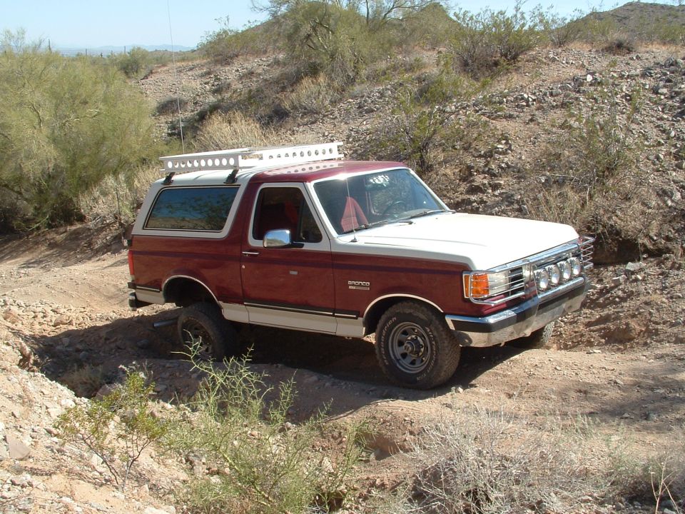 Two-tone red and white Ford Bronco with a roof rack driving on a dirt trail