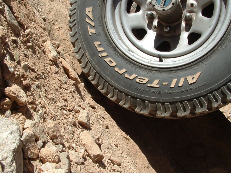 Close-up of a BFGoodrich All-Terrain tire on a silver wheel, partially obscured by dirt and rocks