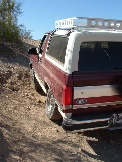 1990 Ford Bronco - A red and white 1990 Ford Bronco navigates a dusty, off-road trail under a clear blue sky.