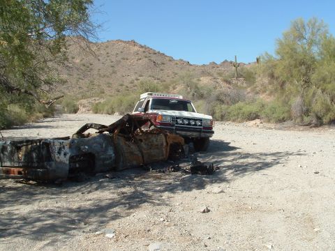 1990 Ford Bronco - A burnt-out vehicle lies in a dry wash, with a support vehicle parked behind it under a clear blue sky.