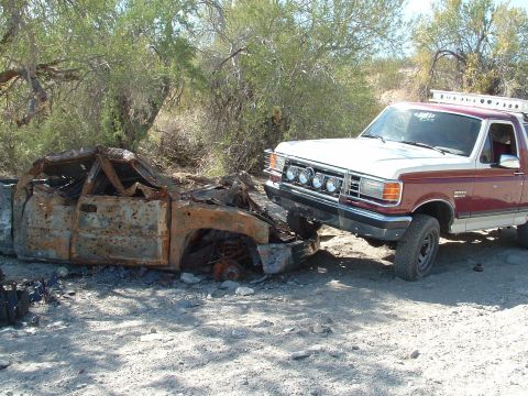 1990 Ford Bronco - A 1990 Ford Bronco is parked beside a destroyed vehicle in a desert landscape.