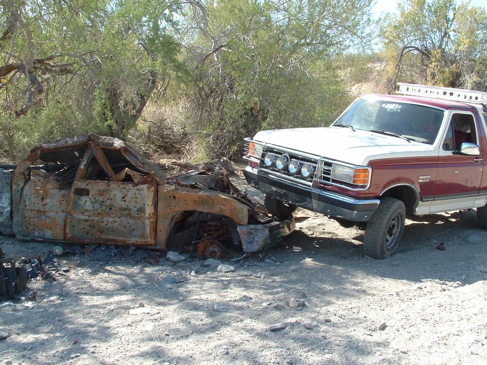 1990 Ford Bronco next to a heavily damaged, rusted car. The Bronco has a light bar on the front