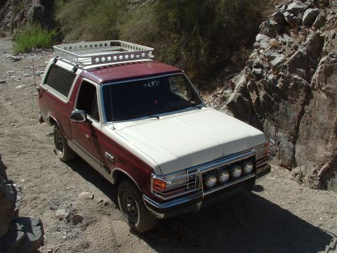 1990 Ford Bronco - A two-tone red and white 1990 Ford Bronco equipped with a roof rack navigates a rocky, off-road trail.