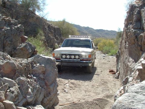 1990 Ford Bronco - A light-colored Ford F-Series truck navigates a rocky, off-road trail under a clear sky.