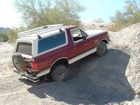 1990 Ford Bronco - This 1990 Ford Bronco navigates a rough dirt terrain, equipped with a roof rack and a rear-mounted spare tire.