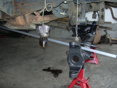 1991 Ford Bronco - Vehicle undercarriage components are visible, supported by red jack stands, with a bag of fluid suspended and a spill on the concrete floor.