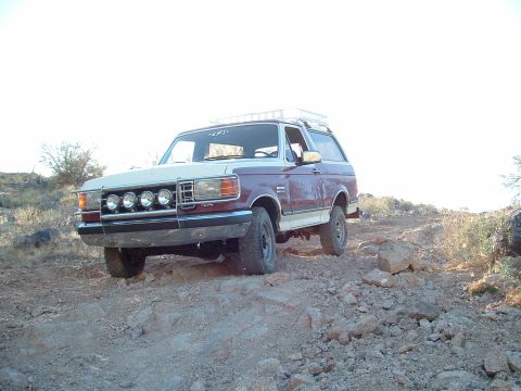 1990 Ford Bronco - A two-tone maroon and white 1990 Ford Bronco navigates a rocky, off-road trail under a bright sky.