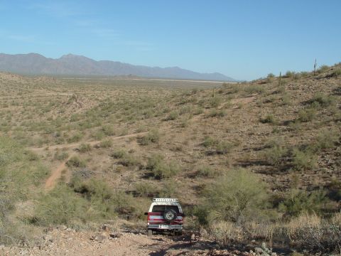 1990 Ford Bronco - A red and white SUV, identified as a 1990 Ford Bronco, drives on a dirt road through a desert landscape.