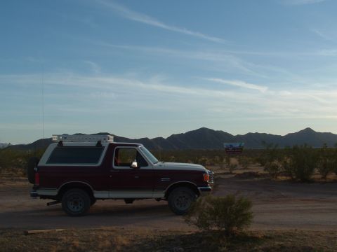 1990 Ford Bronco - A two-tone Ford Bronco sits idle on a dusty desert road under a vast blue sky.