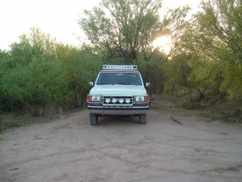 1990 Ford Bronco - A white SUV with a brush guard and roof rack is parked on a dirt road surrounded by trees.