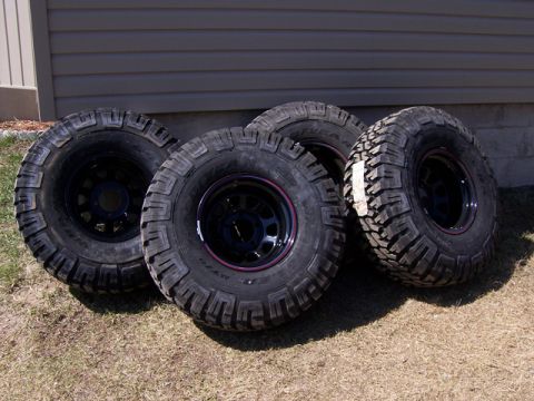 1996 Ford Bronco - Four Goodyear MTR tires with aggressive tread patterns are displayed on black rims, resting on dry grass in front of a building.