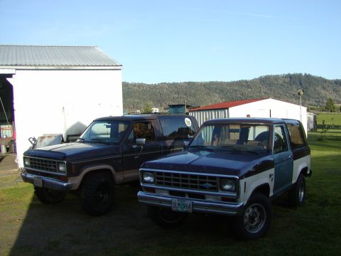 1987 Ford Bronco II - Two Ford Bronco II SUVs are parked in a yard with buildings and hills in the background.