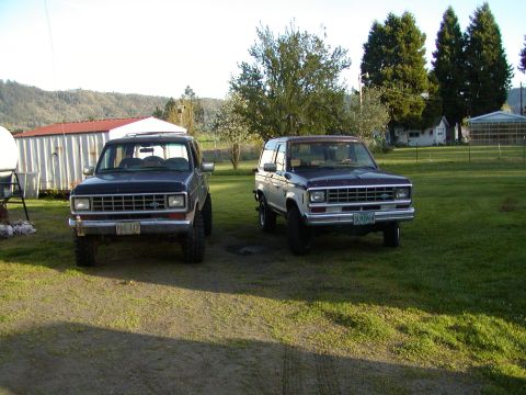 1987 Ford Bronco II - Two Ford Bronco II SUVs are parked in a grassy area with trees and buildings in the background.