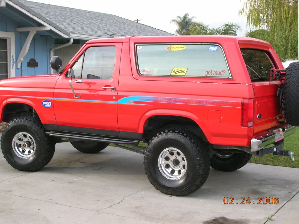 Lifted red two-door Ford Bronco on mud tires parked in a driveway by a blue house