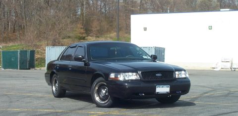 1999 Ford Crown Victoria - A black Ford Crown Victoria sedan photographed from the front-right three-quarter angle in an empty parking lot, with black steel wheels, t…