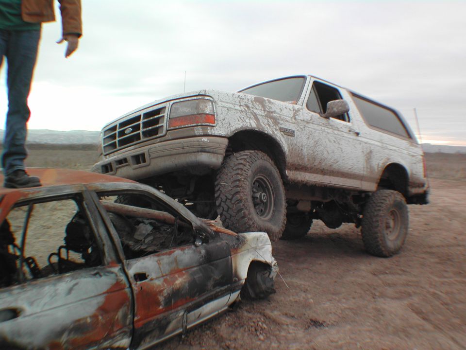 1996 Ford Bronco on a dirt road, driving over a burnt car. A person stands on the burnt car