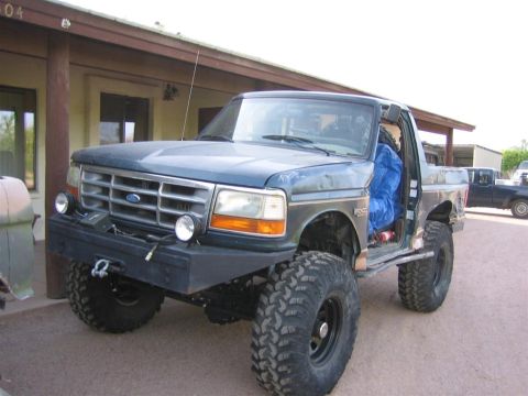 1994 Ford Bronco - A modified Ford Bronco with oversized tires and a custom bumper sits outside a building, with another truck visible in the background.