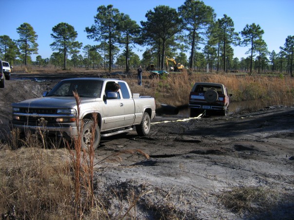 Silver Chevy pickup towing a stuck tan SUV out of a muddy pit off-road