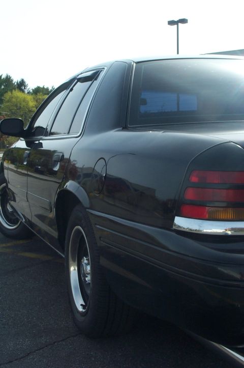 1999 Ford Crown Victoria - Low rear three-quarter view of a black full-size sedan parked on asphalt, showing the vertical red taillight with amber turn section, chrom…