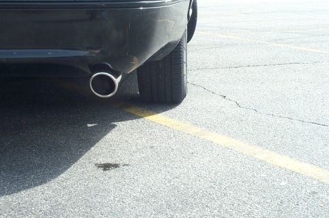 1999 Ford Crown Victoria - Close-up of the rear corner of a dark gray/black sedan, showing a chrome exhaust tip below the bumper and the rear tire, parked on cracked…