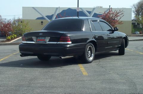 1999 Ford Crown Victoria - A black Ford Crown Victoria full-size sedan viewed from the rear three-quarter angle, parked in an empty asphalt lot with tinted windows, c…