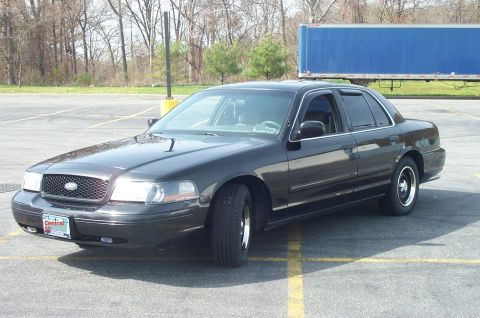 1999 Ford Crown Victoria - A dark brown/black Ford Crown Victoria full-size sedan parked in an asphalt lot, shown from a front three-quarter angle with black steel wh…
