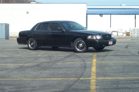 1999 Ford Crown Victoria - A black Ford Crown Victoria sedan shown in a 3/4 front side view, parked in an empty asphalt lot with tire skid marks. It has black steel w…