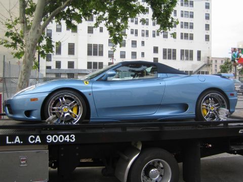 1990 Ford Bronco - light blue Ferrari convertible with chrome five-spoke wheels and yellow brake calipers sits strapped to a flatbed truck on an urban street.