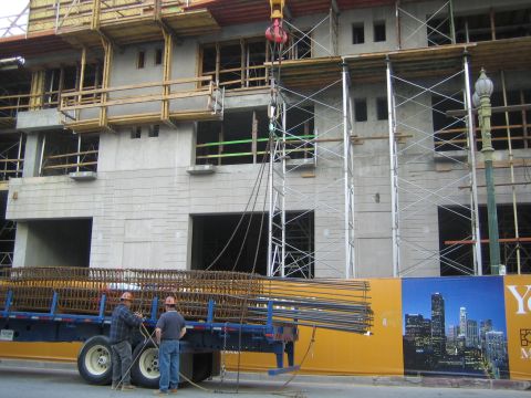1990 Ford Bronco - Two workers stand by a flatbed loaded with bundled rebar in front of a multi-story concrete building under construction with scaffolding.