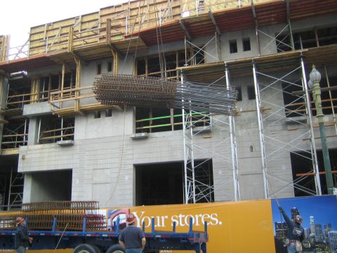 1990 Ford Bronco - long bundled rebar cage hangs near a concrete building under construction, with an orange 'Your stores' hoarding below.