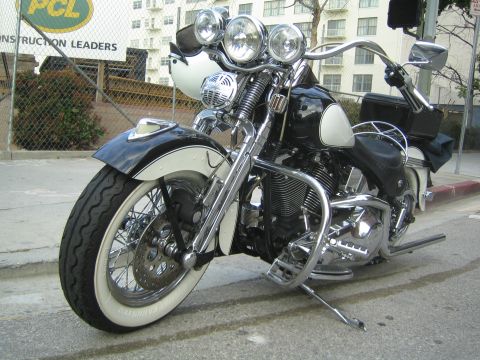 1990 Ford Bronco - black-and-white cruiser motorcycle with triple headlights and whitewall tires stands on its kickstand near a PCL Construction fence.
