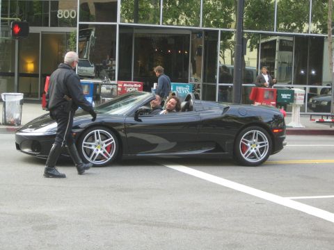 1990 Ford Bronco - black Ferrari convertible carrying two occupants waits on a city street while a bald man in black walks across in front of it.