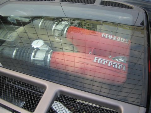 1990 Ford Bronco - View through a mesh engine cover of a red-finned engine bay with chrome intake clamps and silver 'Ferrari' lettering.