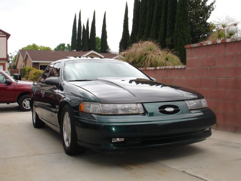 1993 Ford Taurus - A dark green Ford Taurus with a modified hood is parked in front of a brick wall and residential buildings.