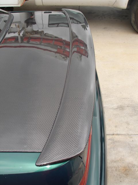 1993 Ford Taurus - The trunk of a dark green car features a carbon fiber spoiler, with reflections of a building visible on the surface.