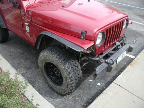 1998 Jeep TJ - Front corner of a red Jeep Wrangler showing off-road tires and a damaged aftermarket bumper.