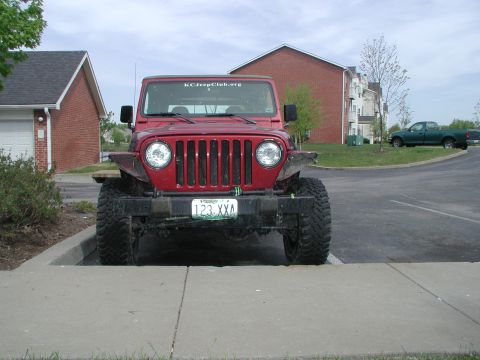 1998 Jeep TJ - A red Jeep Wrangler is parked in front of a building, with a green pickup truck visible in the background.