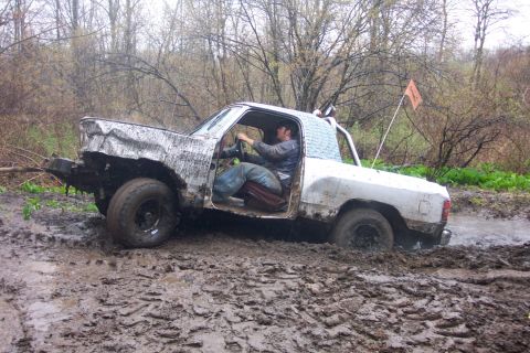 1978 Ford Bronco - A driver navigates a heavily damaged white pickup truck through a muddy off-road course.