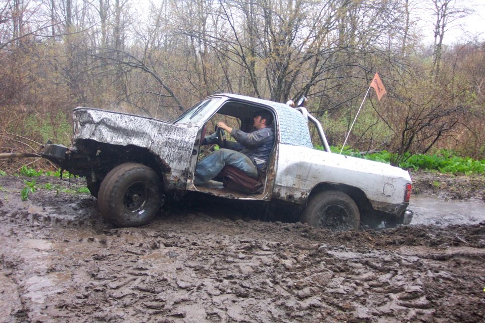 Mud-splattered white pickup truck with driver, stuck in thick mud with trees in background
