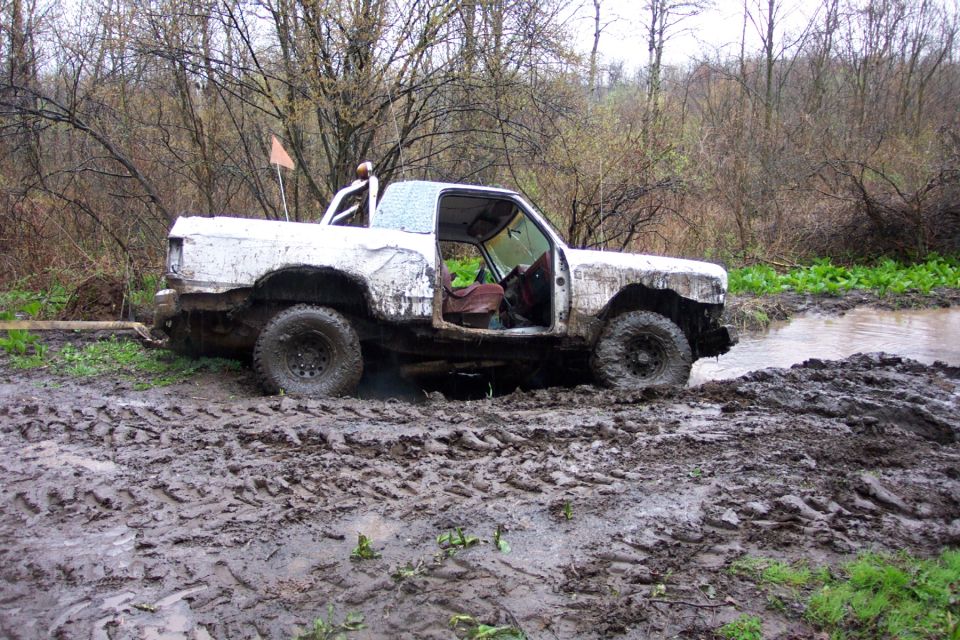 White off-road vehicle with doors open, stuck in deep mud and water. Trees and brush in background