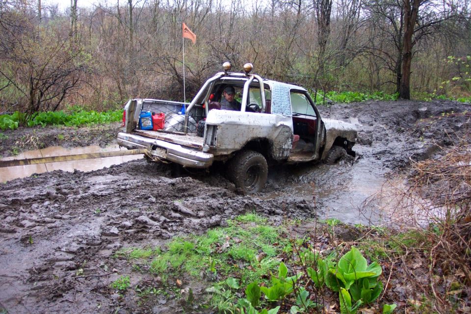Silver off-road vehicle with roll cage stuck in deep mud, orange flag on antenna, gas cans in bed