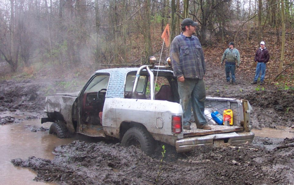 Pickup truck stuck deep in mud with people standing around. One person is in the truck bed