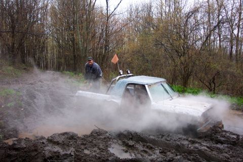 1978 Ford Bronco - A man watches as his off-road vehicle gets stuck deep in the mud, creating a large spray.