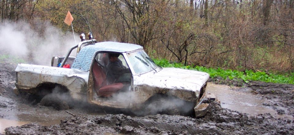 White pickup truck stuck in mud, driver visible, orange flag on roll bar