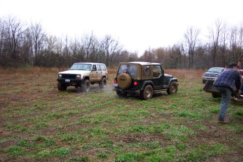 1978 Ford Bronco - Two Jeeps navigate a muddy, grassy field under an overcast sky, with a truck and person visible on the right.