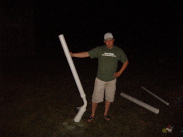 Man in a green t-shirt and shorts holding a large white pipe, standing outdoors at night