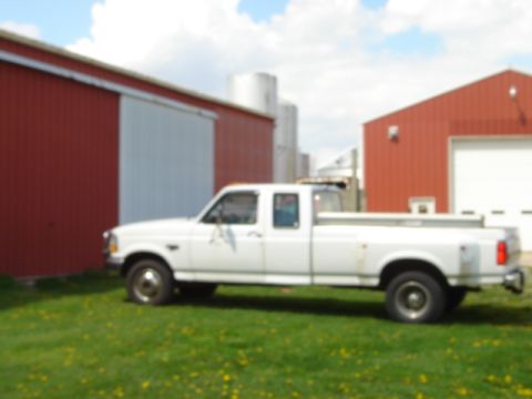 1992 Ford Bronco - A white pickup truck is parked on a grassy area next to farm buildings under a cloudy sky.