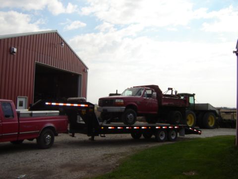 1992 Ford Bronco - Red Ford dump truck loaded onto a trailer, towed by a pickup truck near a red barn.