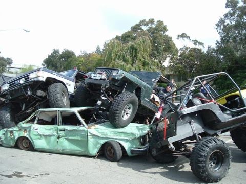 1989 Ford Bronco - Multiple off-road vehicles are piled on top of a crushed green sedan.