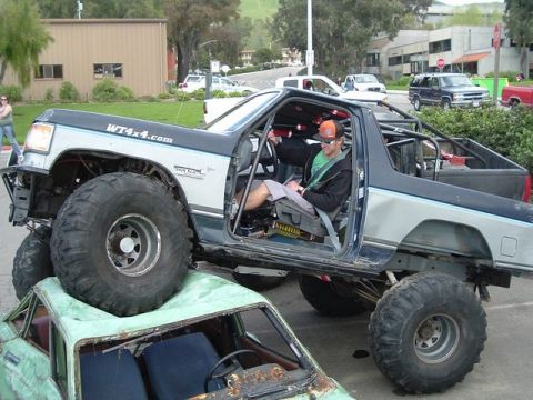 1989 Ford Bronco - A heavily modified off-road truck with oversized tires is shown driving over a car.